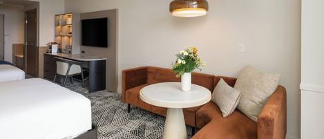 A hotel room with a bed, a sofa, a small round table with a vase of flowers, a desk with a flat-screen TV, and a pendant light.