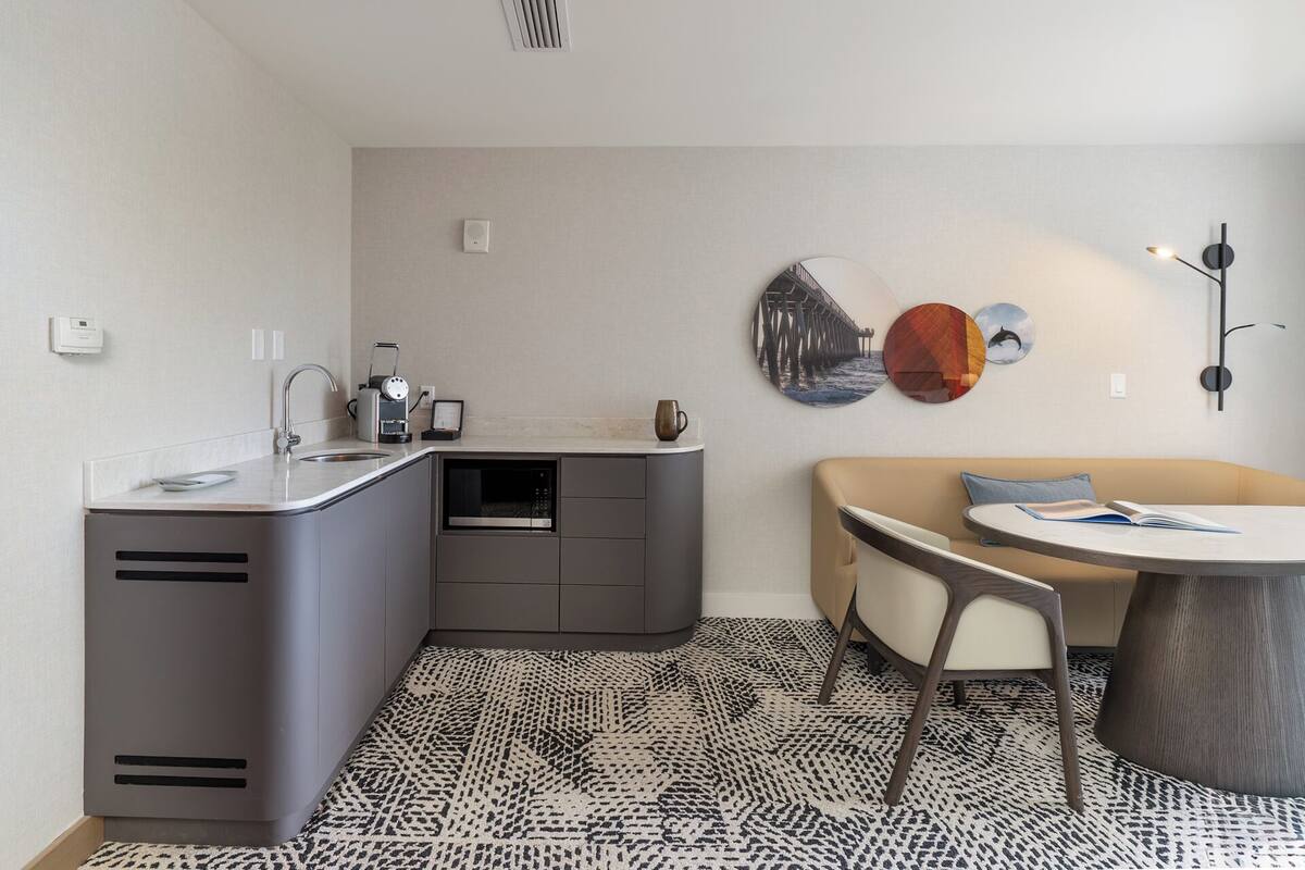 A compact kitchen with a built-in oven, a coffee machine, and a sink. There is a round table with a chair and a sofa with a cushion.