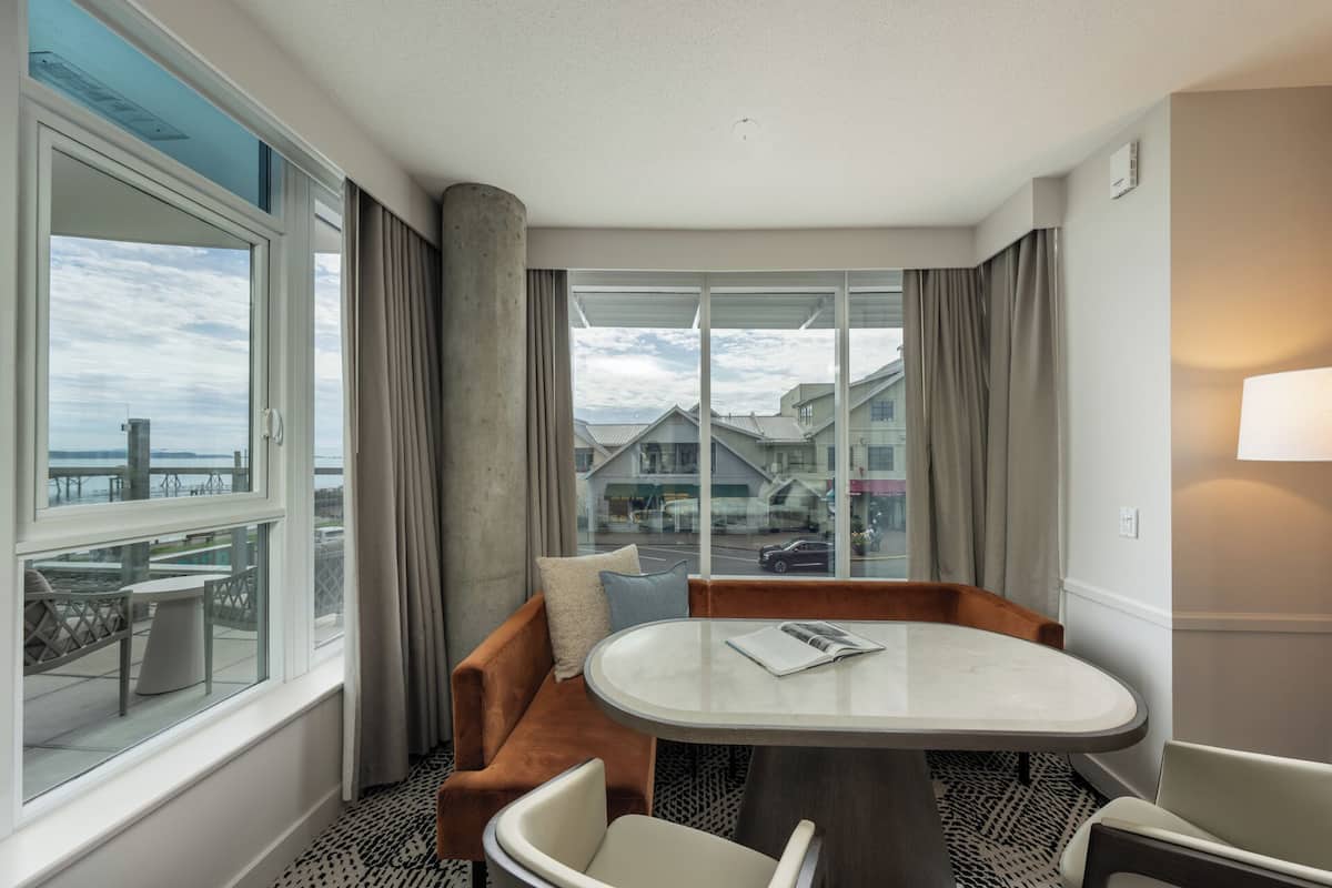 A hotel room with a large window, a round table, a sofa, and a chair.