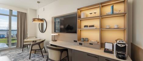 A modern hotel room with a large window, a dining area, a minibar, and a flat-screen TV.
