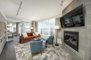 Una habitación de hotel moderna con chimenea, sofá, zona de comedor y ventanas grandes con vistas.