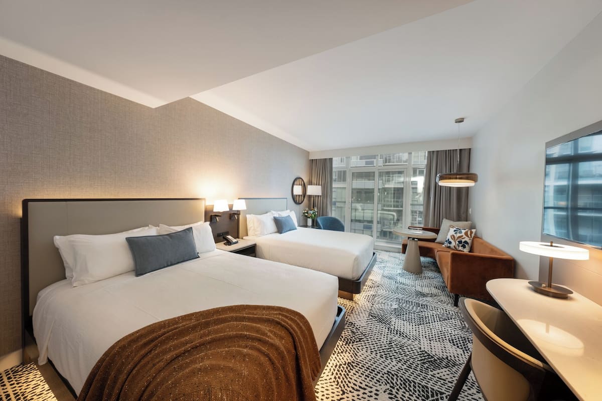 A hotel room with two beds, a desk, a chair, a window with a view, and a patterned rug.