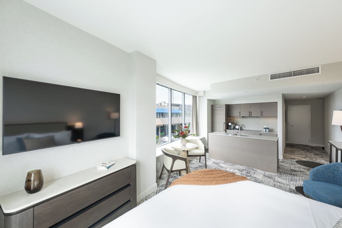 A hotel room with a large bed, a TV mounted on the wall, a small kitchenette, and a seating area with a table and chairs.