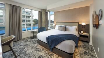 Habitación de hotel con una cama grande, un escritorio, una silla, una mesita de noche y vistas de la ciudad a través de amplios ventanales.
