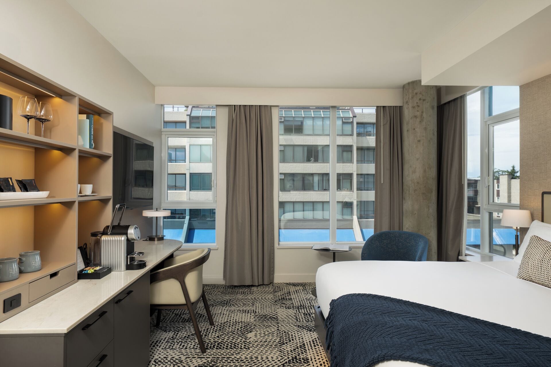 A hotel room with a bed, desk, chair, and a view of a building through the window.