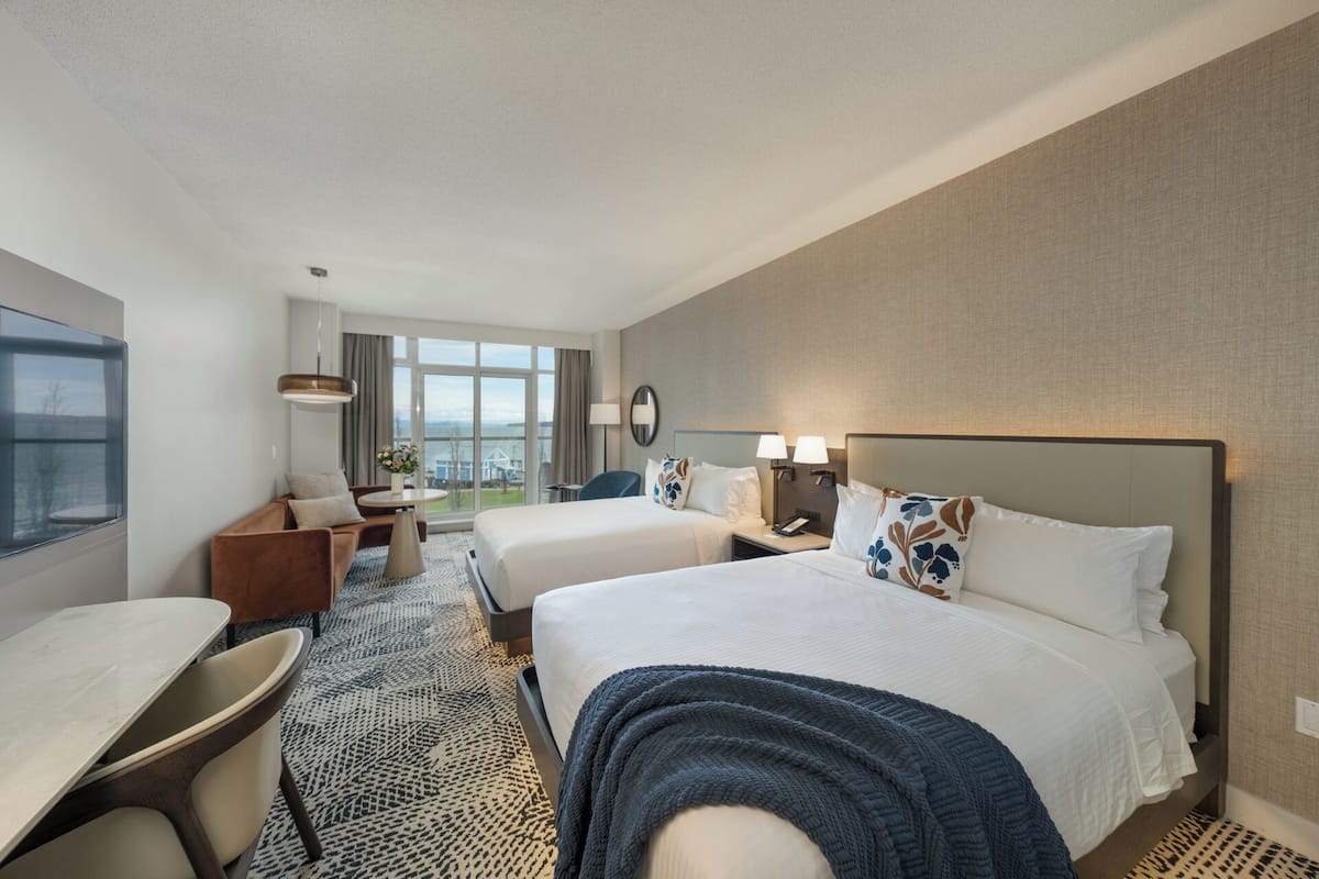A hotel room with two beds, a desk, a chair, a small table, and a large window offering a view.
