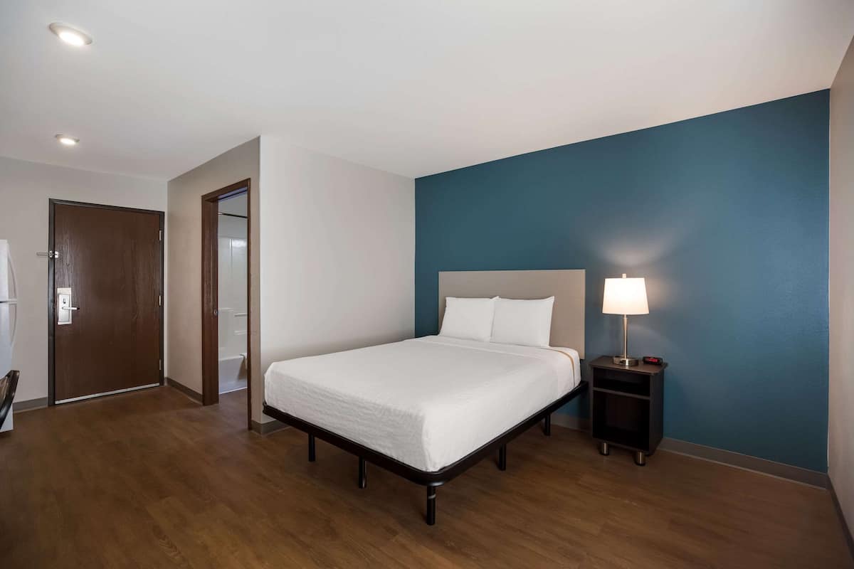 A hotel room with a bed, a nightstand with a lamp, a door, and a bathroom visible through an open door.