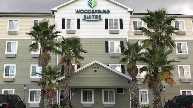 Ein Hotel namens Woodspring Suites mit modernem Design, das ein auffälliges Logo, zahlreiche Fenster und Palmen im Eingangsbereich zeigt.