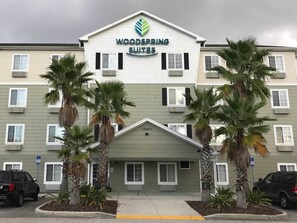 A hotel named Woodspring Suites with a modern design, featuring a prominent logo, multiple windows, and palm trees in the entrance area.