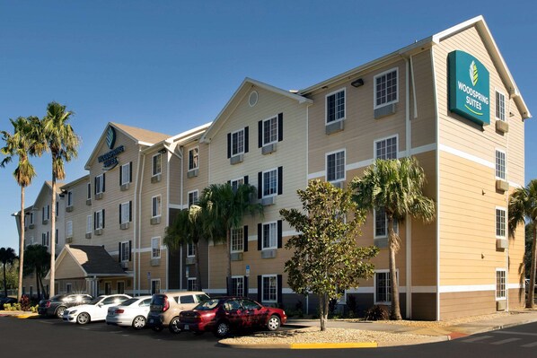 A multi-story hotel with a sign reading "Woodspring Suites." There are several cars parked in front, and palm trees are visible around the property.