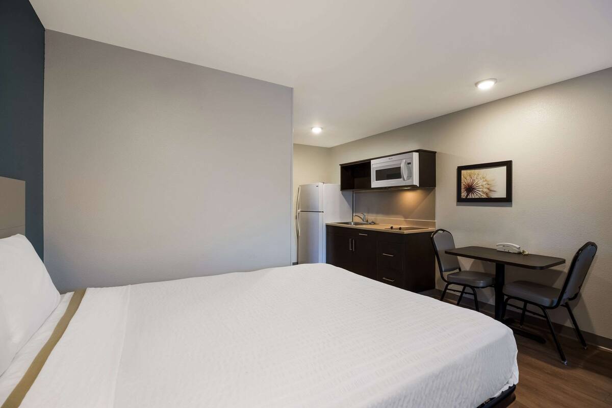 A hotel room with a bed, a small kitchenette with a microwave, refrigerator, and a table with chairs.