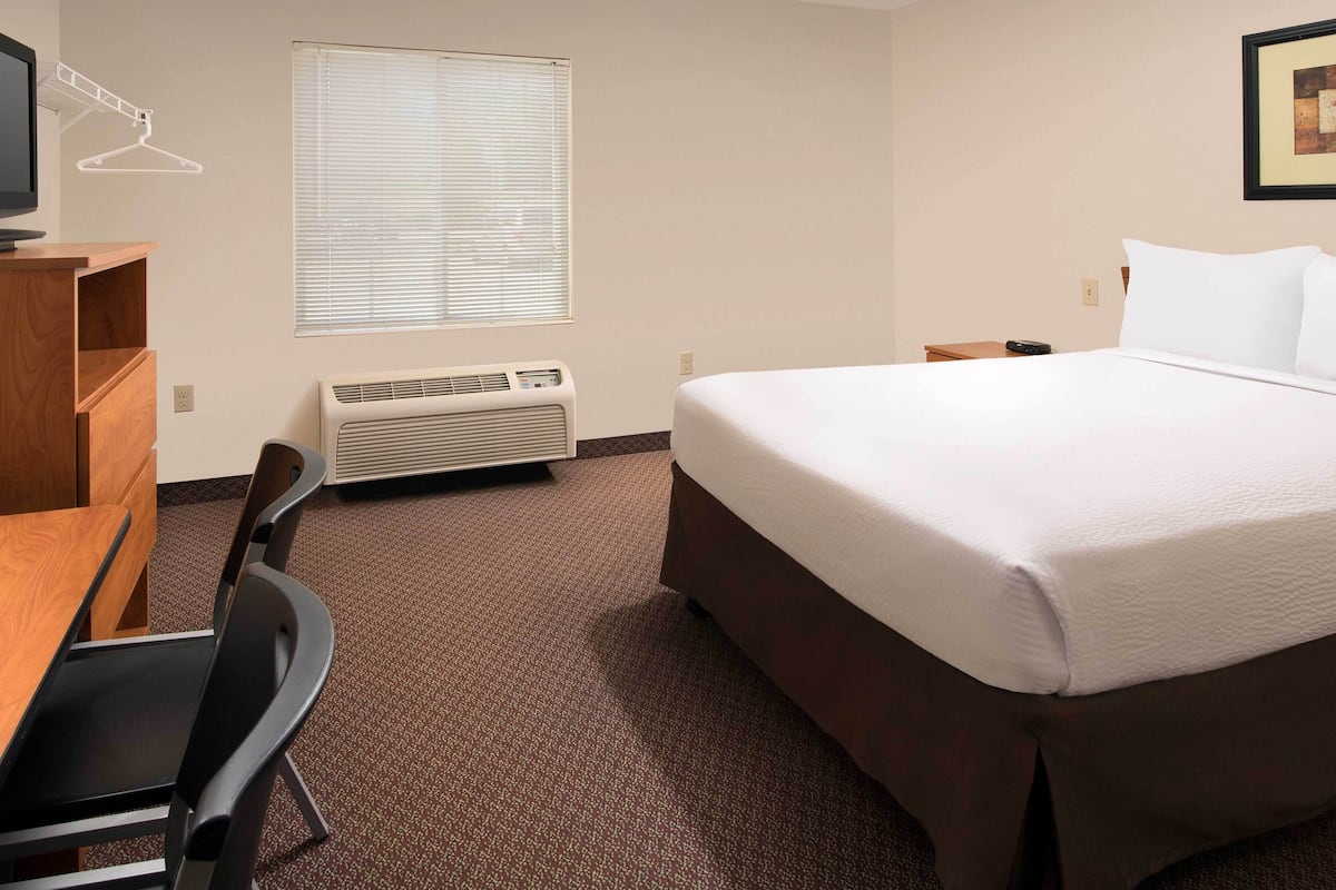 A hotel room with a bed, a desk, a chair, a heater, a window with blinds, and a wall-mounted TV.