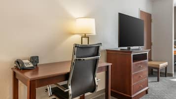 A hotel room with a wooden desk, a black office chair, a lamp, a flat-screen TV, a telephone, and a wooden dresser with drawers.