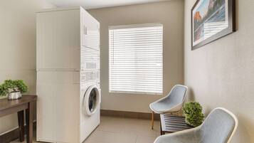 A laundry room with a washing machine, a dryer, a window with blinds, and two chairs.