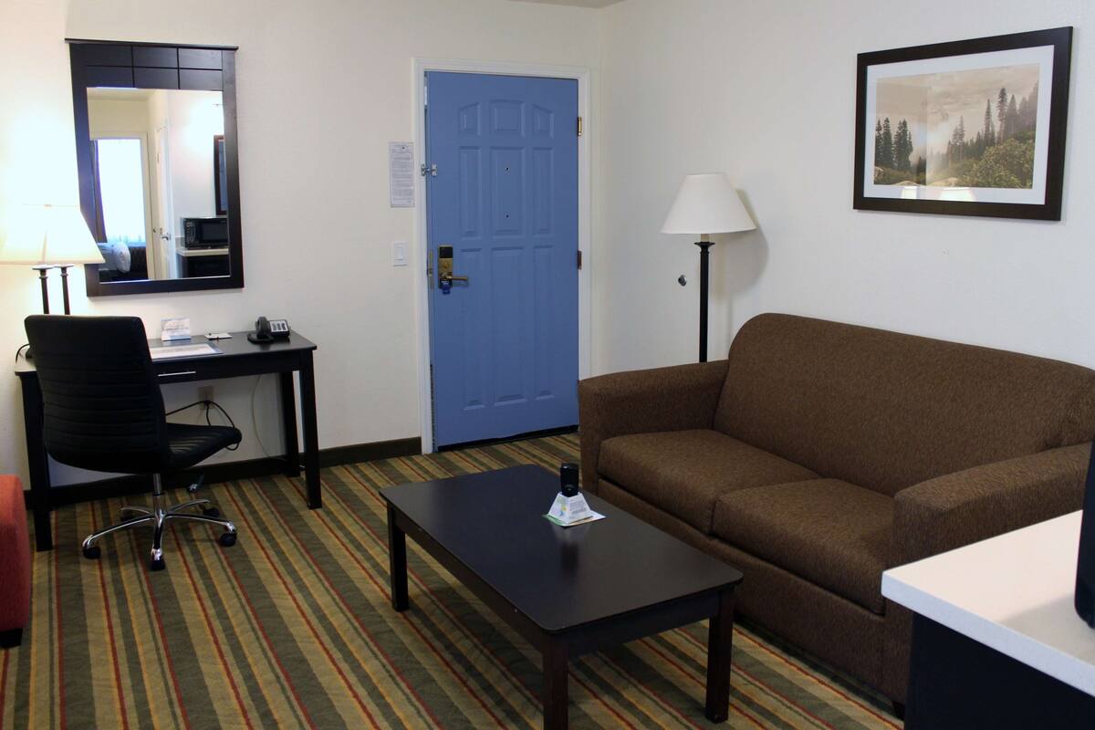 Ein Hotelzimmer mit einem braunen Sofa, einem Schreibtisch mit Stuhl, einem kleinen Tisch, einer Lampe und einem gerahmten Bild an der Wand.