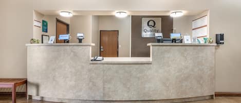 A hotel reception area with a reception desk, a "Quality by Choice Hotels" sign, a reception phone, and a digital kiosk.