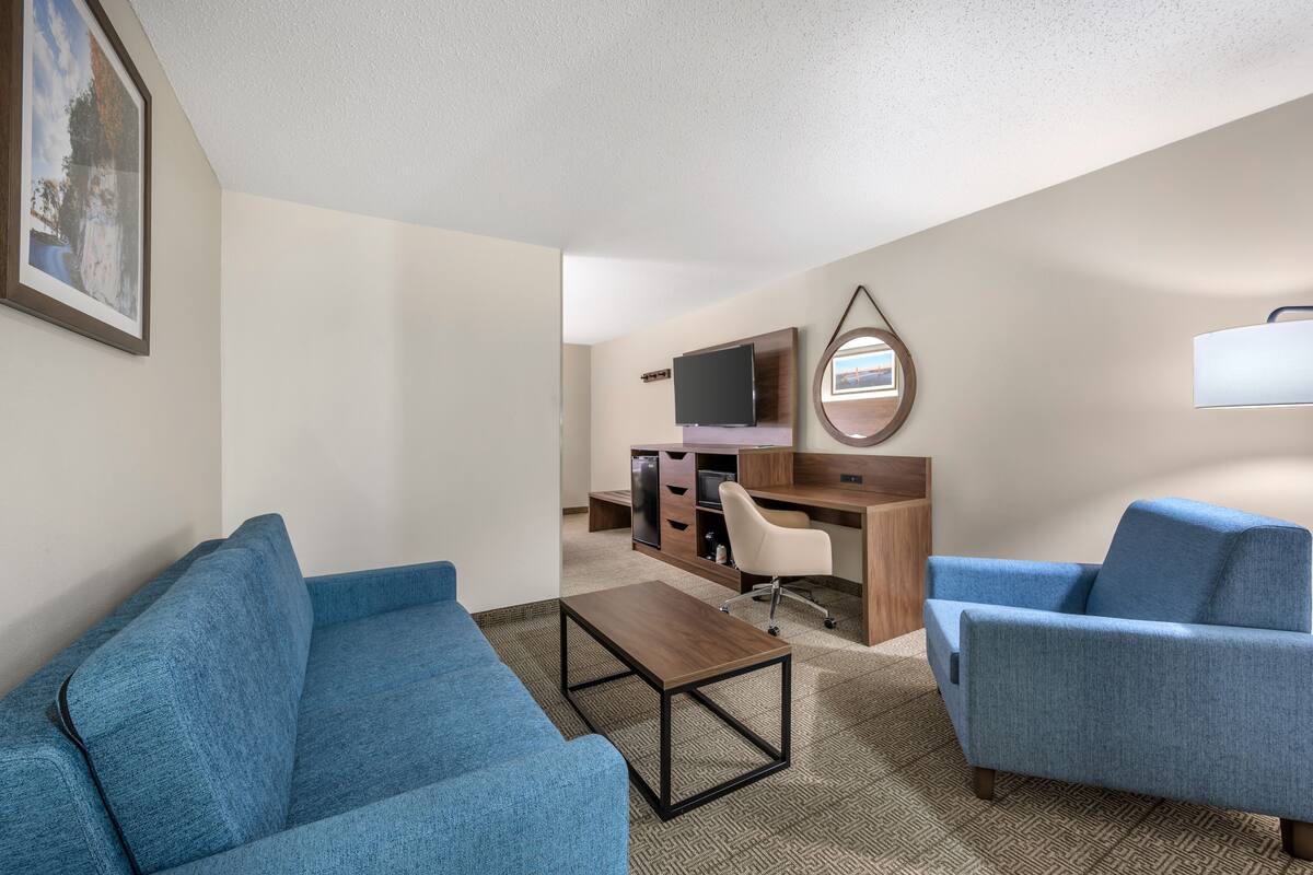 A hotel room with a blue sofa, a wooden coffee table, a desk with a computer and TV, and a small mirror on the wall.