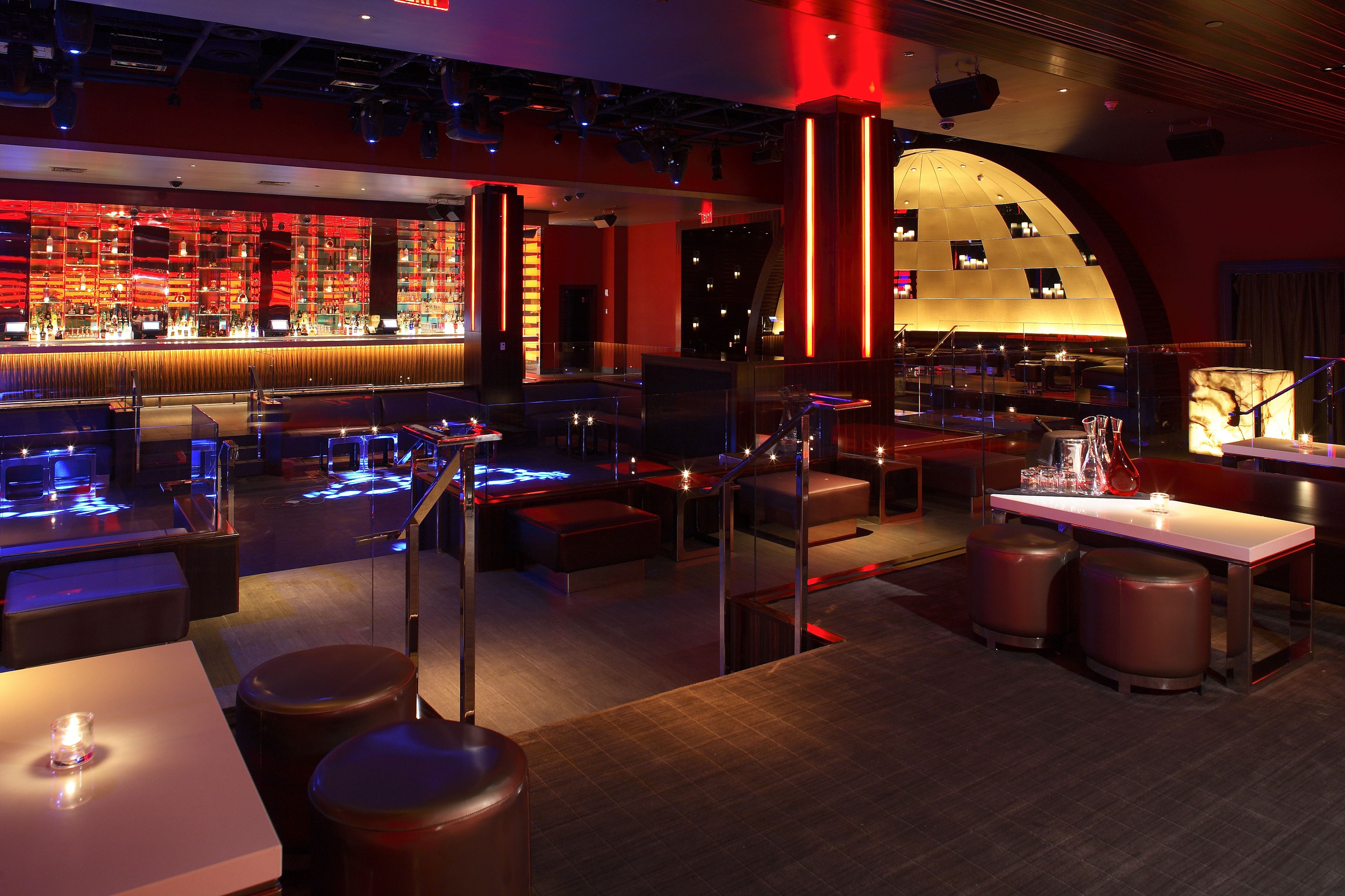 Un área de bar moderna con una barra iluminada en rojo, asientos con taburetes y un escenario para espectáculos.