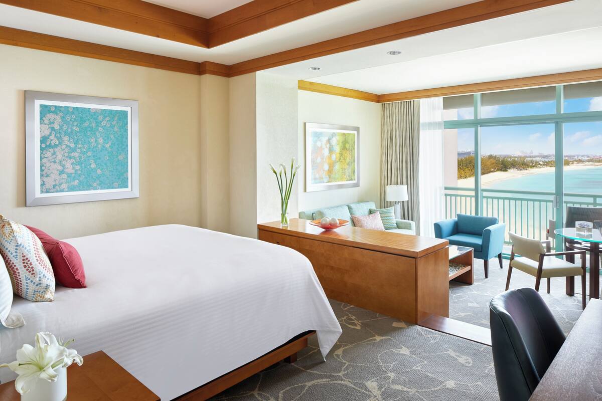 A hotel room with a large bed, a desk with a chair, a sofa, and a balcony with a view of the beach.