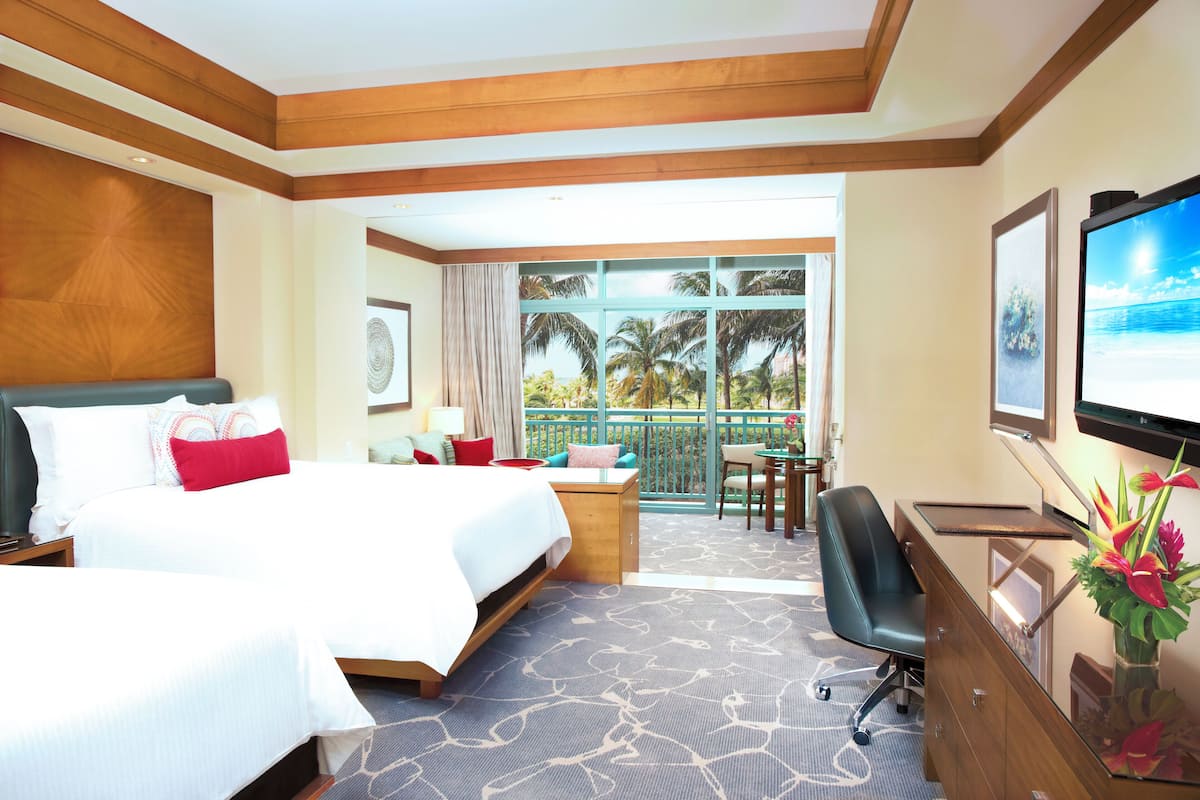A hotel room with two beds, a desk, a chair, a balcony with a view of palm trees, and a TV on the wall.