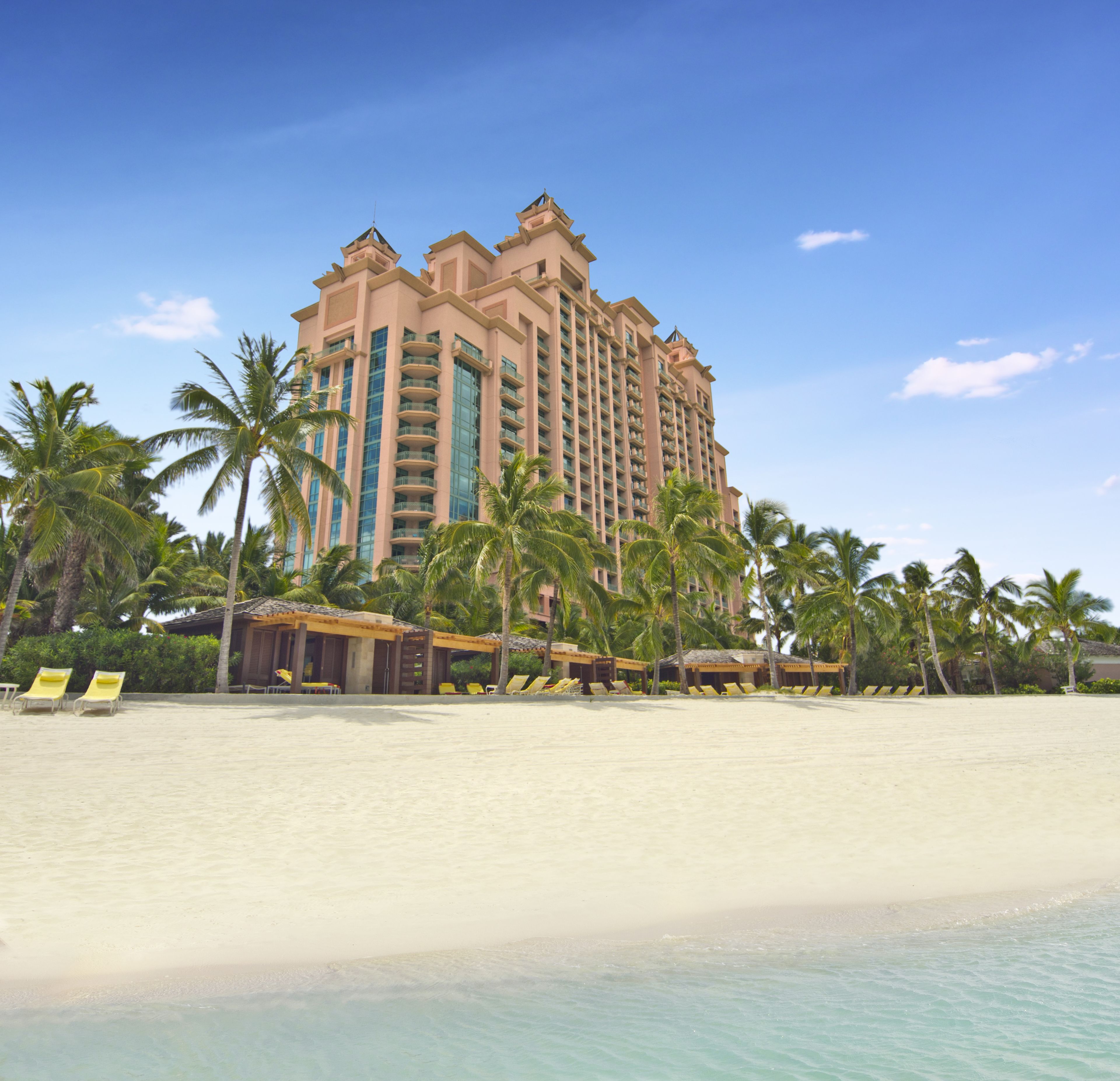 Un hotel frente al mar con varios pisos, palmeras y una playa de arena.