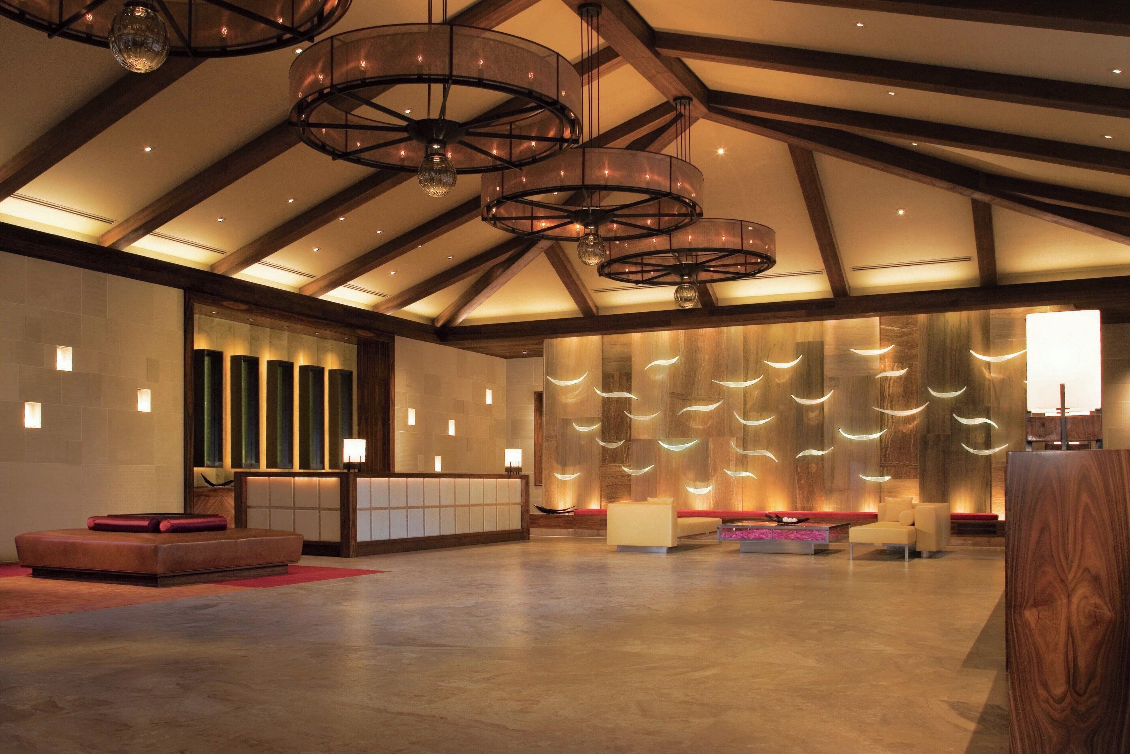 A spacious lobby with a high ceiling, wooden beams, and modern lighting fixtures. There are seating areas with cushions and a reception desk.