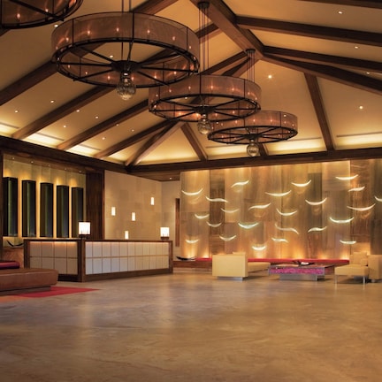A spacious lobby with a high ceiling, wooden beams, and modern lighting fixtures. There are seating areas with cushions and a reception desk.