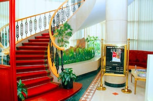 Una majestuosa escalera con peldaños rojos y barandilla de cristal que conduce a un área de recepción con una planta grande en maceta y un cartel con información del hotel.