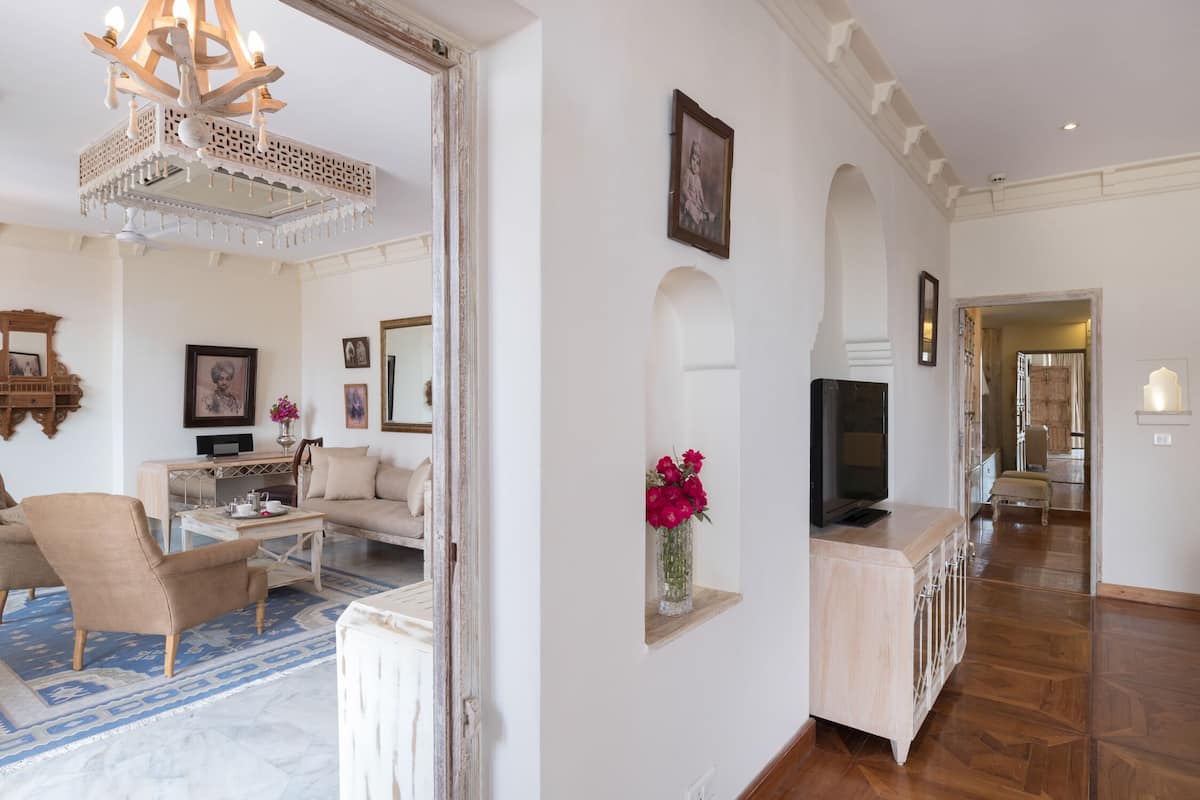 A spacious living room with a sofa, armchairs, a coffee table, and a TV on a wooden cabinet. There is a chandelier, a mirror, and a vase with flowers on the wall.