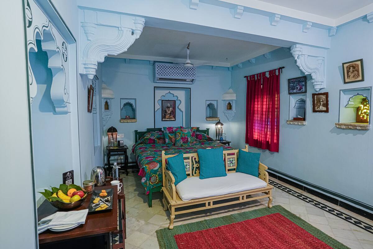 A room with a bed, a small table with a fruit bowl, a red curtain, and decorative wall elements.