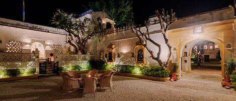 Un patio exterior con zona de estar, una fuente y un edificio con ventanas de arco y un balcón.