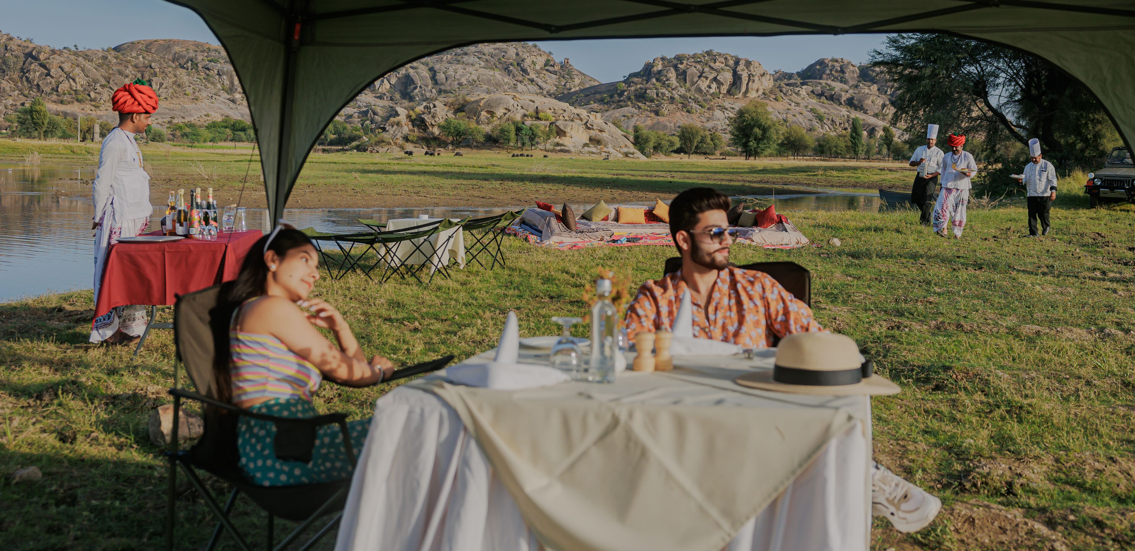 A tented dining area with a table set for two, a person in traditional attire, and a scenic view of a lake and mountains in the background.