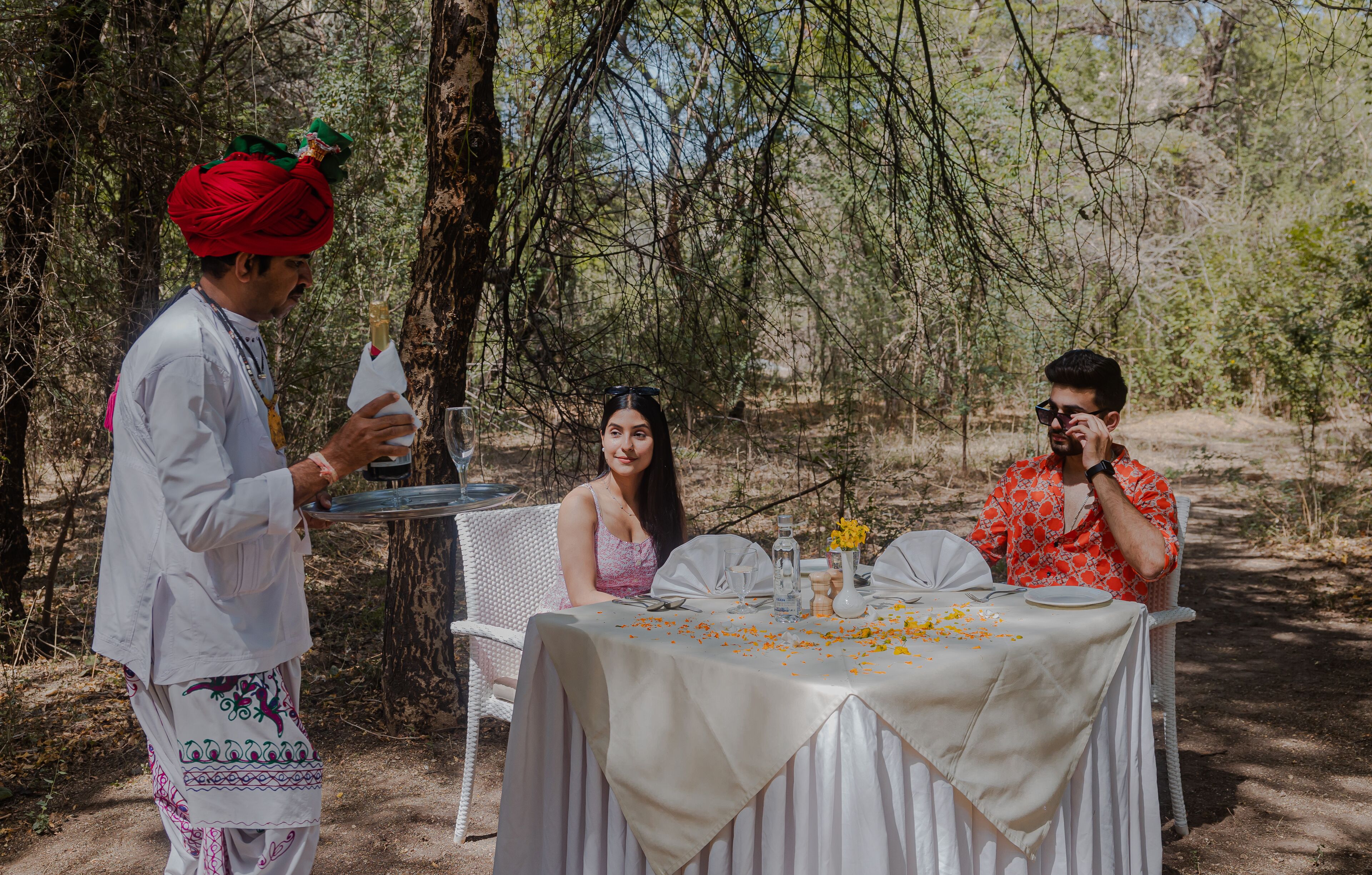 A dining table set outdoors with a white tablecloth, surrounded by trees.