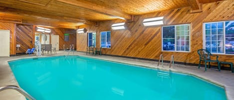 An indoor swimming pool with wooden walls and ceiling, clear windows, and a few chairs around the pool.