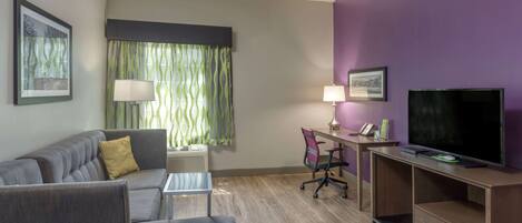 A modern hotel room with a grey sofa, a small table, a desk with a chair, a TV, and a purple accent wall.