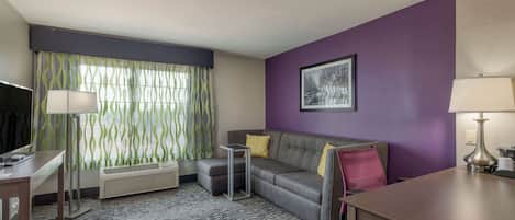 Habitación de hotel con sofá gris, una mesita, un escritorio, una lámpara de pie, un cuadro en la pared y una ventana con cortinas.
