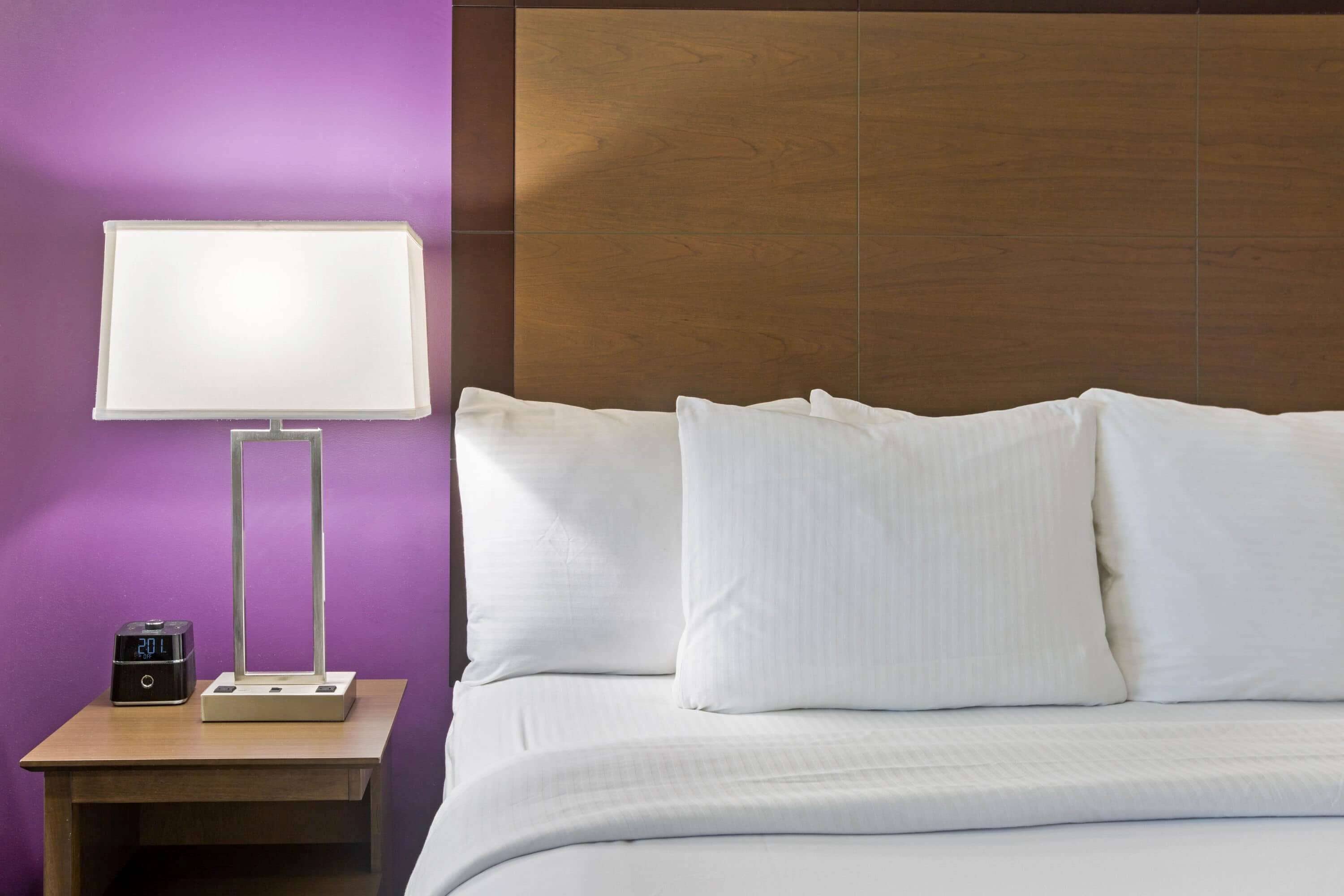 A neatly made bed with white linens and pillows, a bedside table with a lamp and a digital clock, and a wooden headboard.