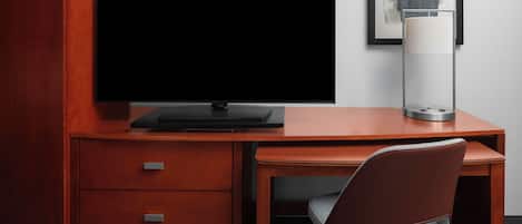 A modern hotel room with a wooden desk, a flat-screen TV, a lamp, and a framed artwork on the wall.