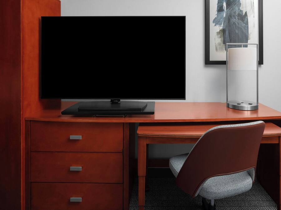 A modern hotel room with a wooden desk, a flat-screen TV, a lamp, and a framed artwork on the wall.
