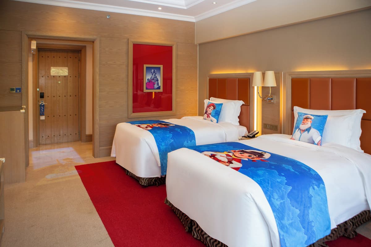 A hotel room with two beds, a red carpet, a wooden door, and a red-framed picture on the wall.
