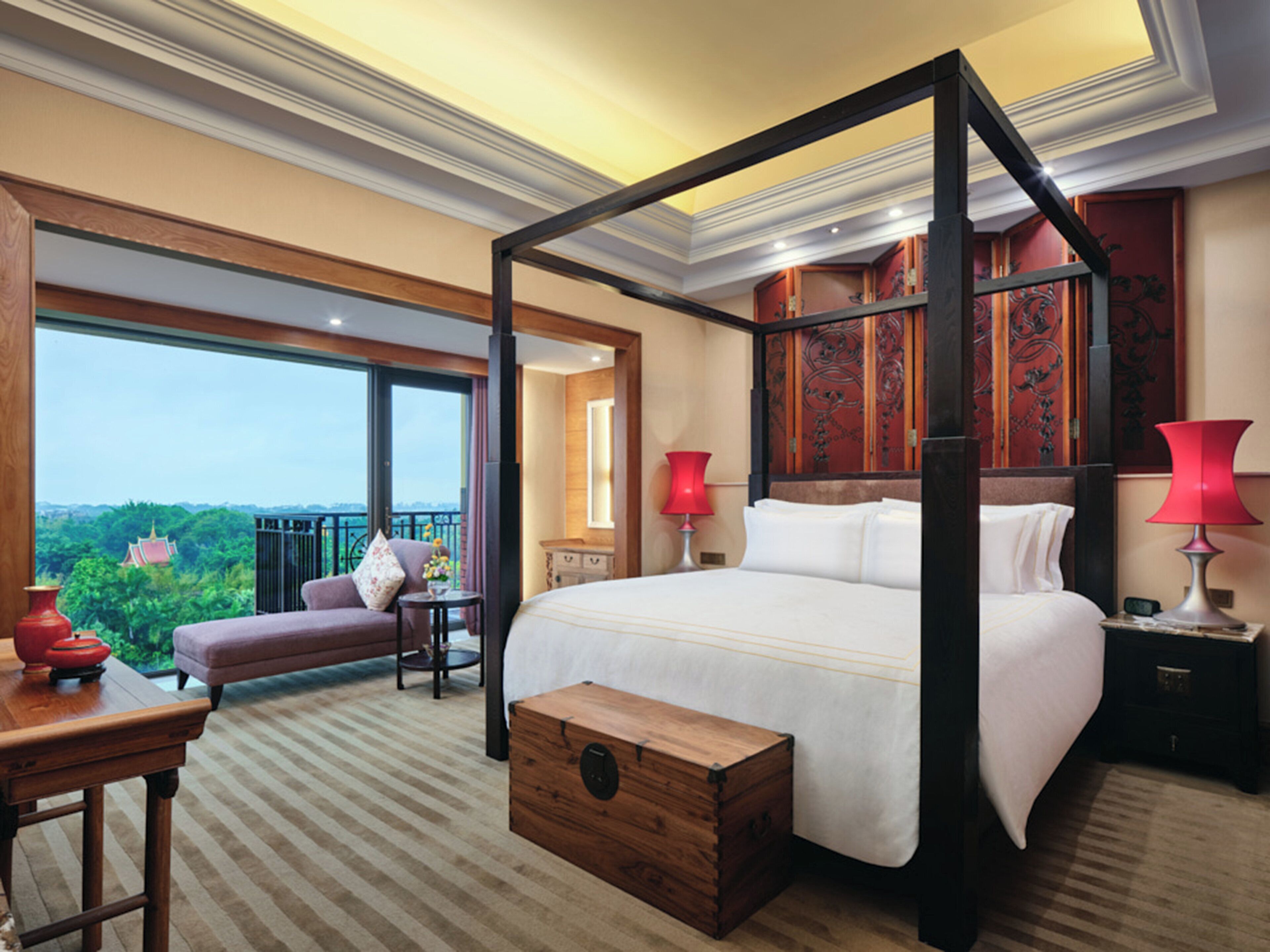 A spacious bedroom with a four-poster bed, a wooden chest, a nightstand, and a red lamp.