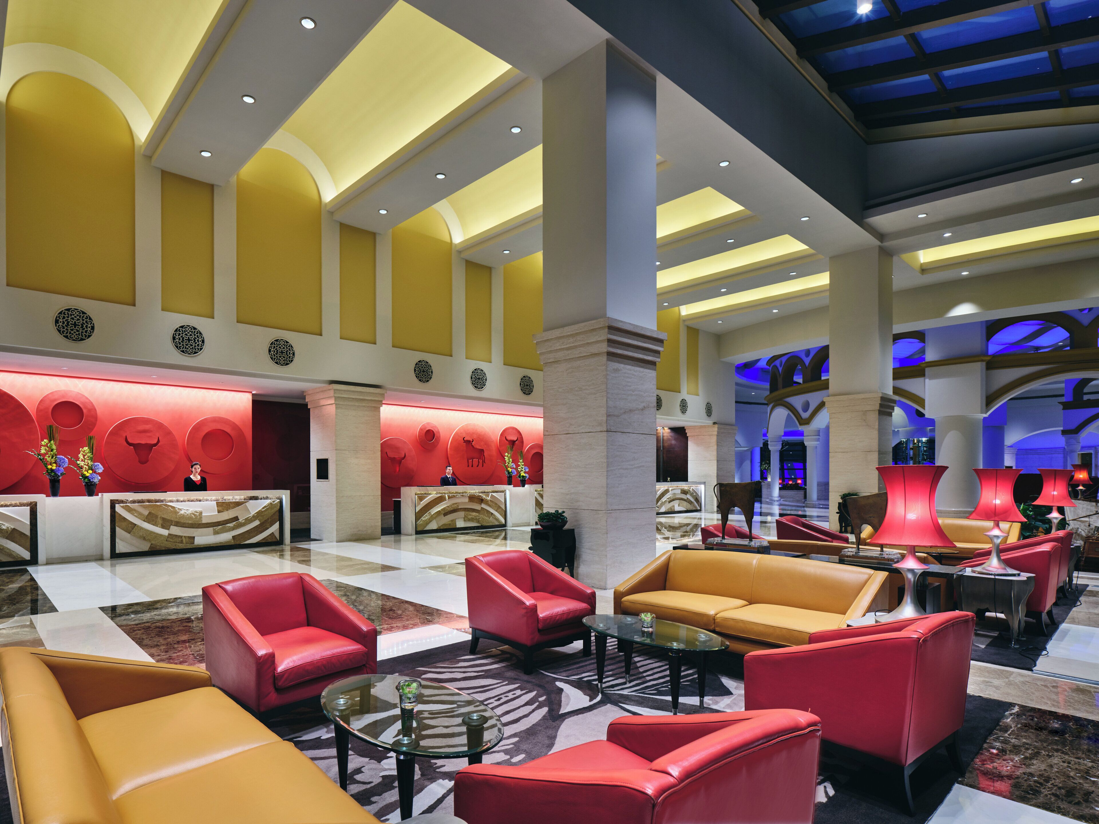 Un vestíbulo de hotel con mostrador de recepción, asientos rojos y amarillos, y un diseño de techo moderno.