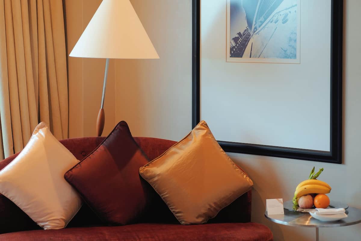 A red sofa with multiple cushions, a standing lamp, a framed picture on the wall, and a small table with fruits.