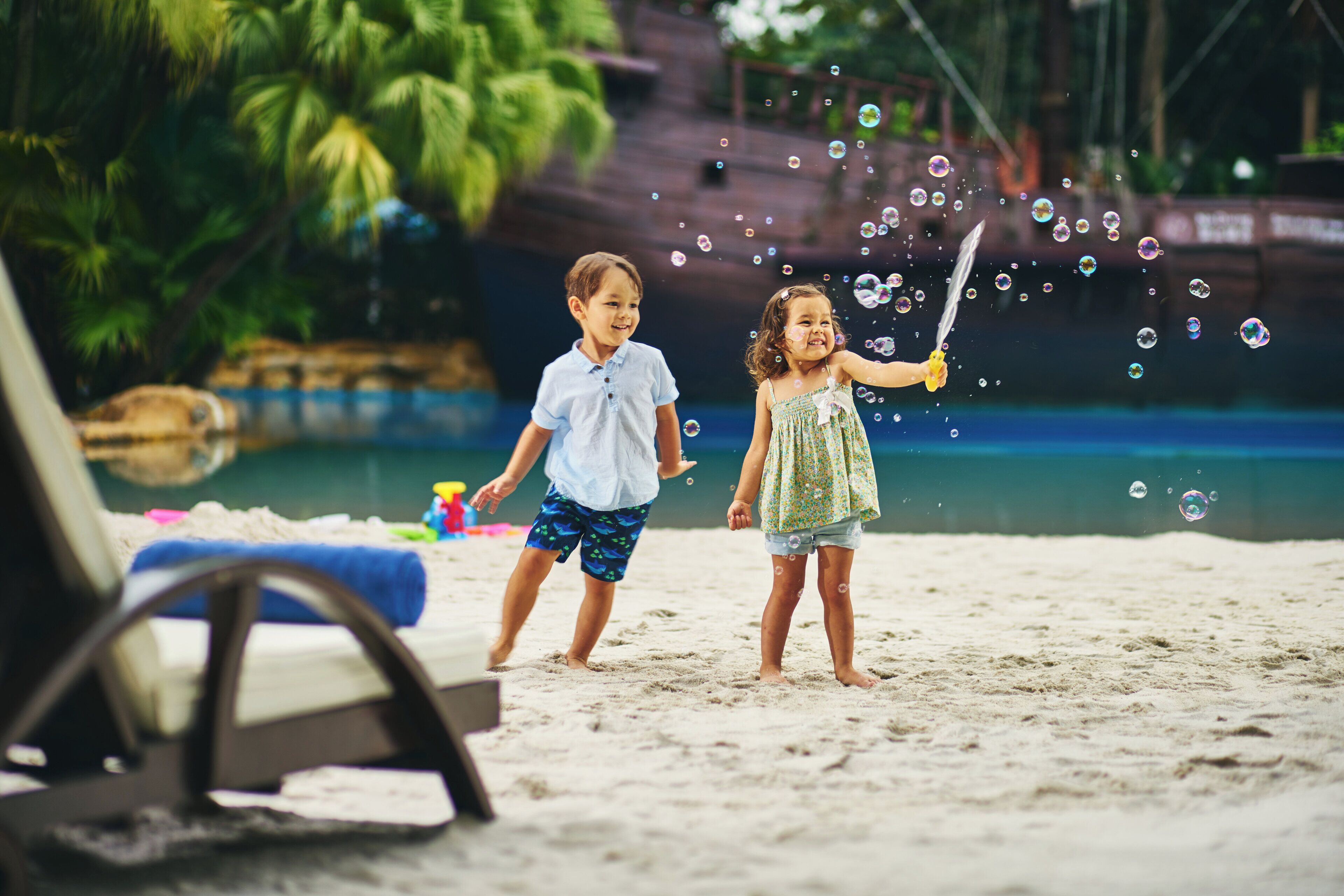 Two children playing with bubbles on a sandy beach with a pool and palm trees in the background.