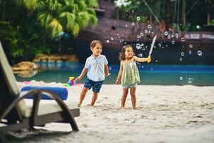 Zwei Kinder spielen mit Seifenblasen an einem sandigen Strand, im Hintergrund ein Pool und Palmen.