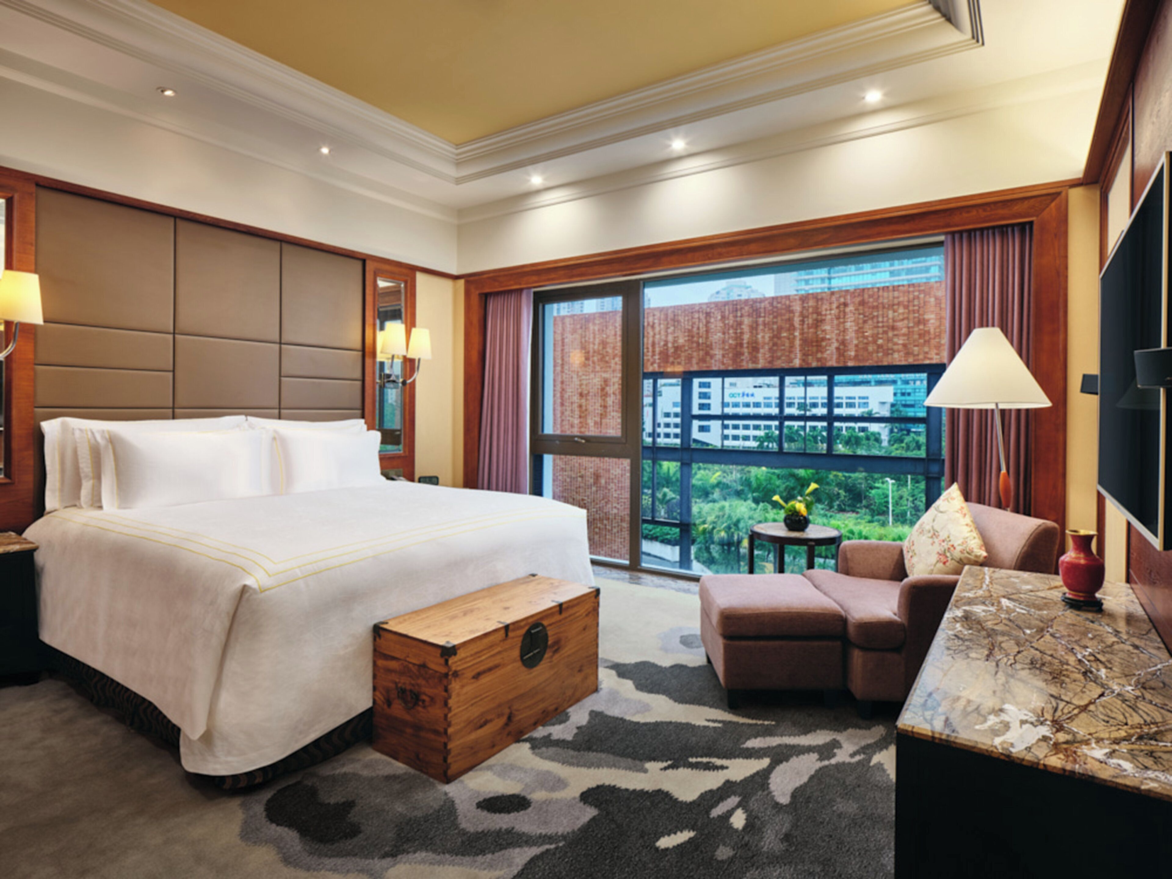 A hotel room with a large bed, a wooden chest, a sofa, a small table, and a large window offering a city view.