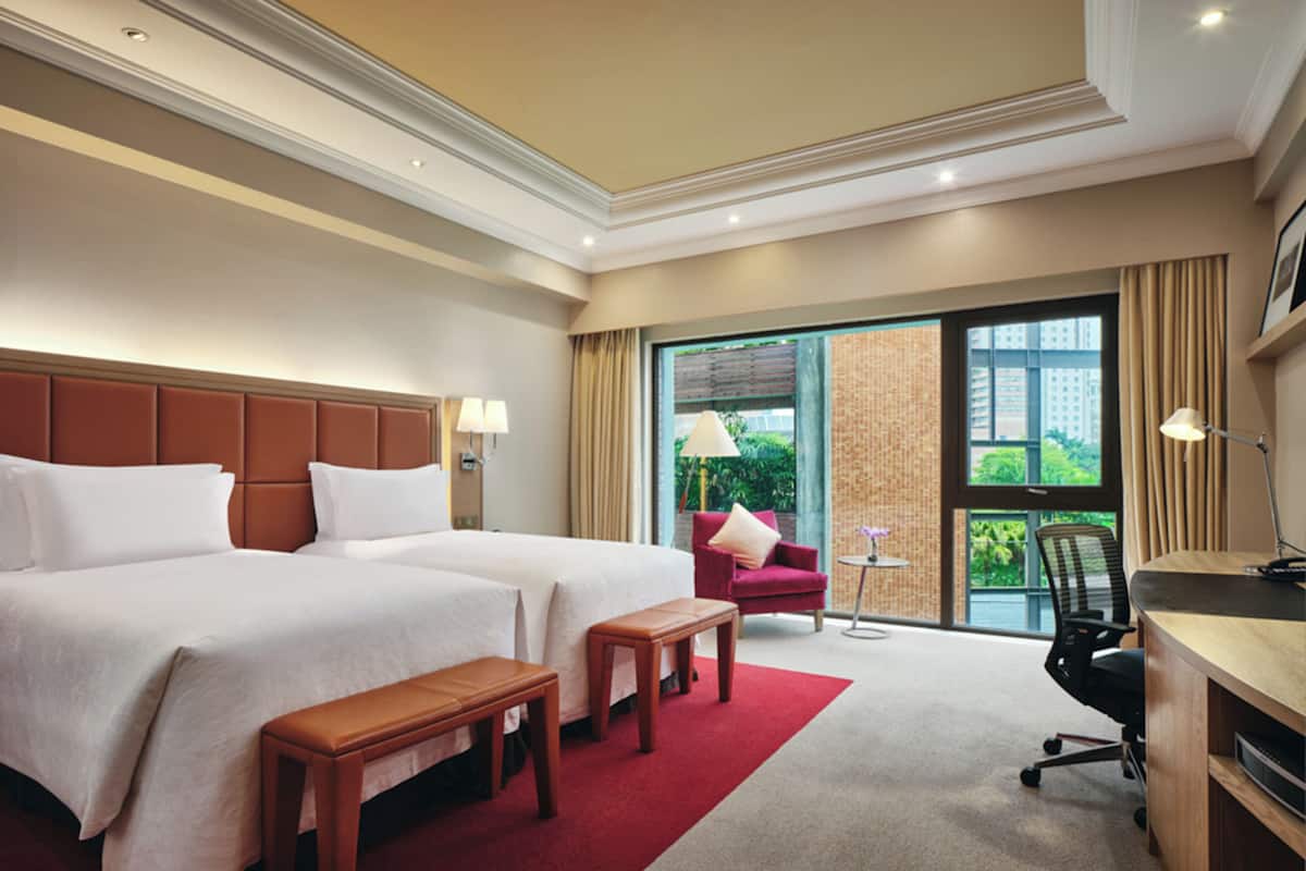 A hotel room with two beds, a desk, a chair, a small table, and a view of greenery outside.