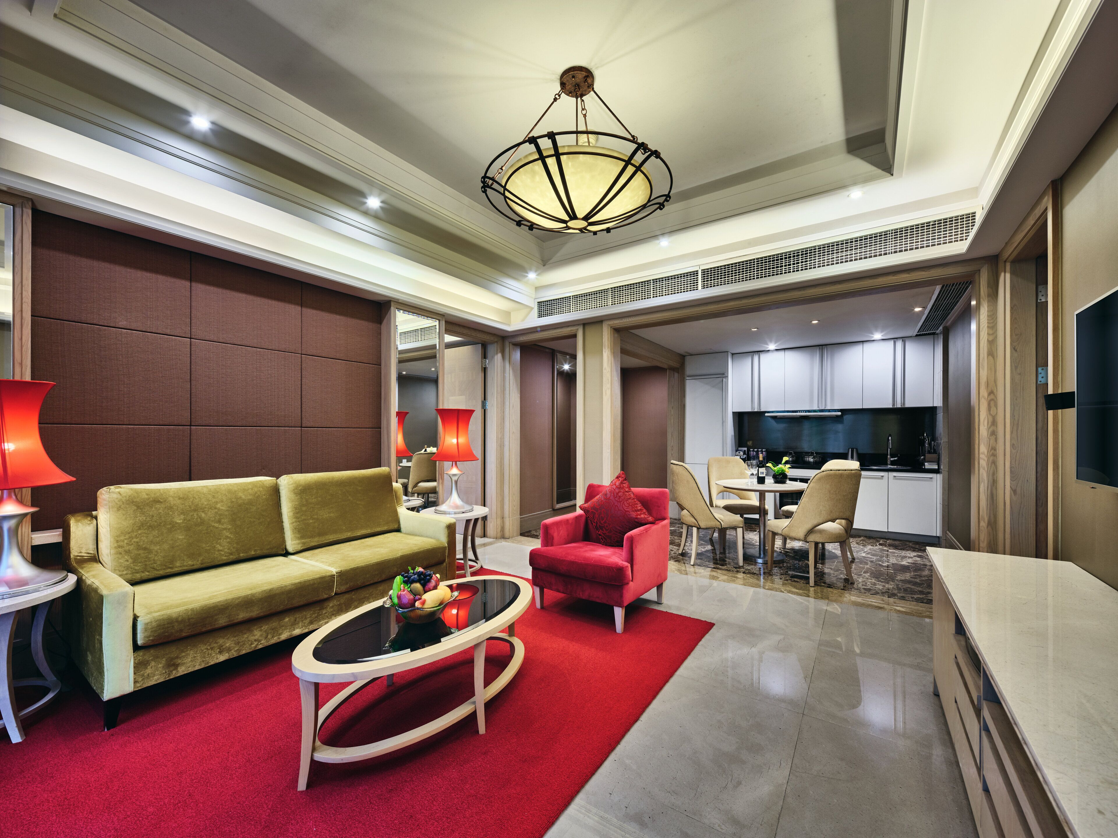 A modern hotel room with a sofa, armchair, coffee table, and a dining area with a TV.
