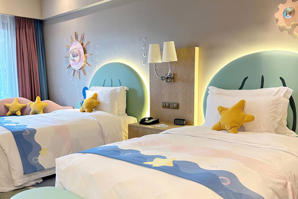A hotel room with two beds, a bench, a nightstand, and a wall clock.