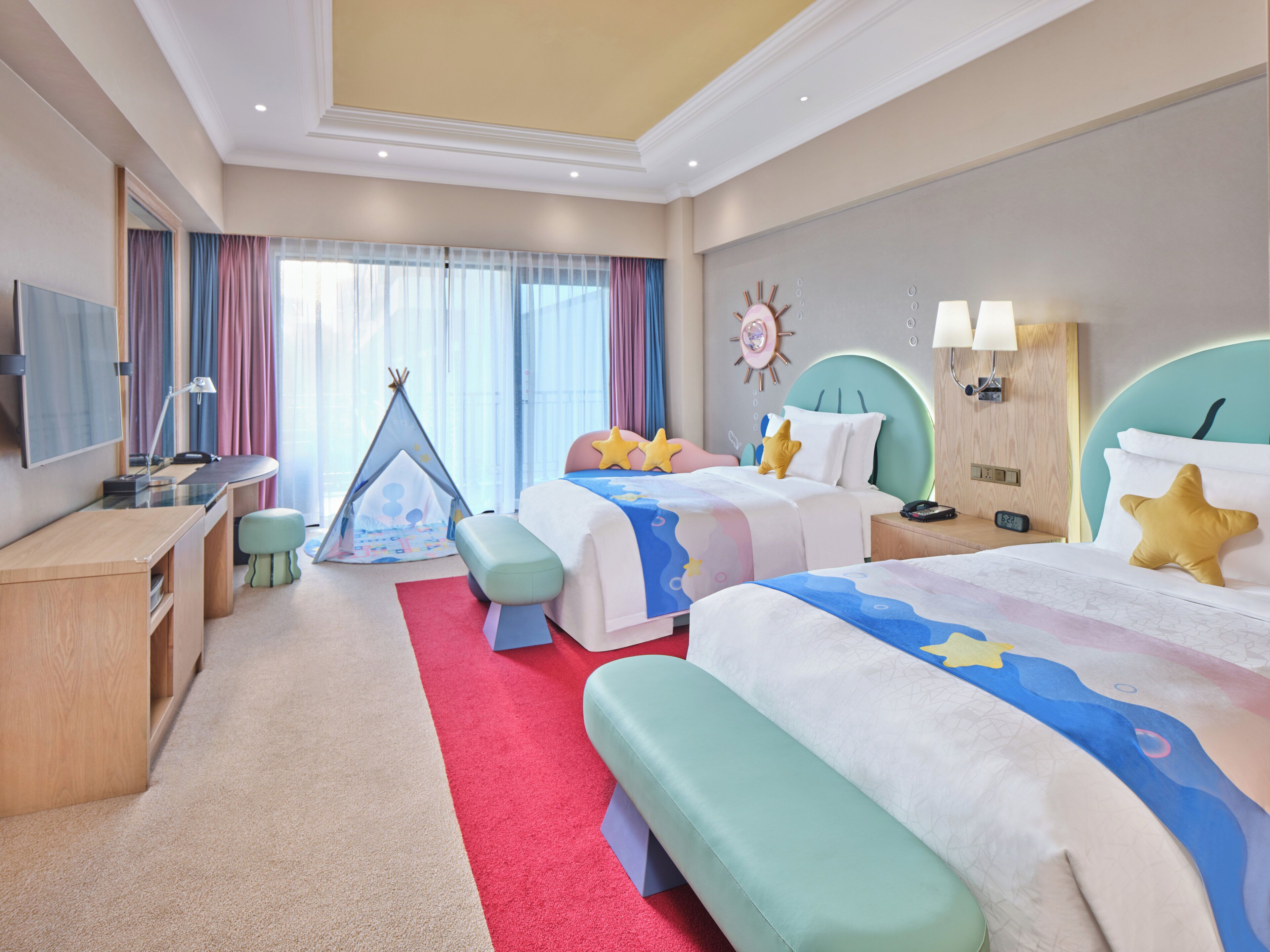 Quarto de hotel com duas camas, uma escrivaninha, uma cadeira, uma tenda infantil e uma janela grande com cortinas.